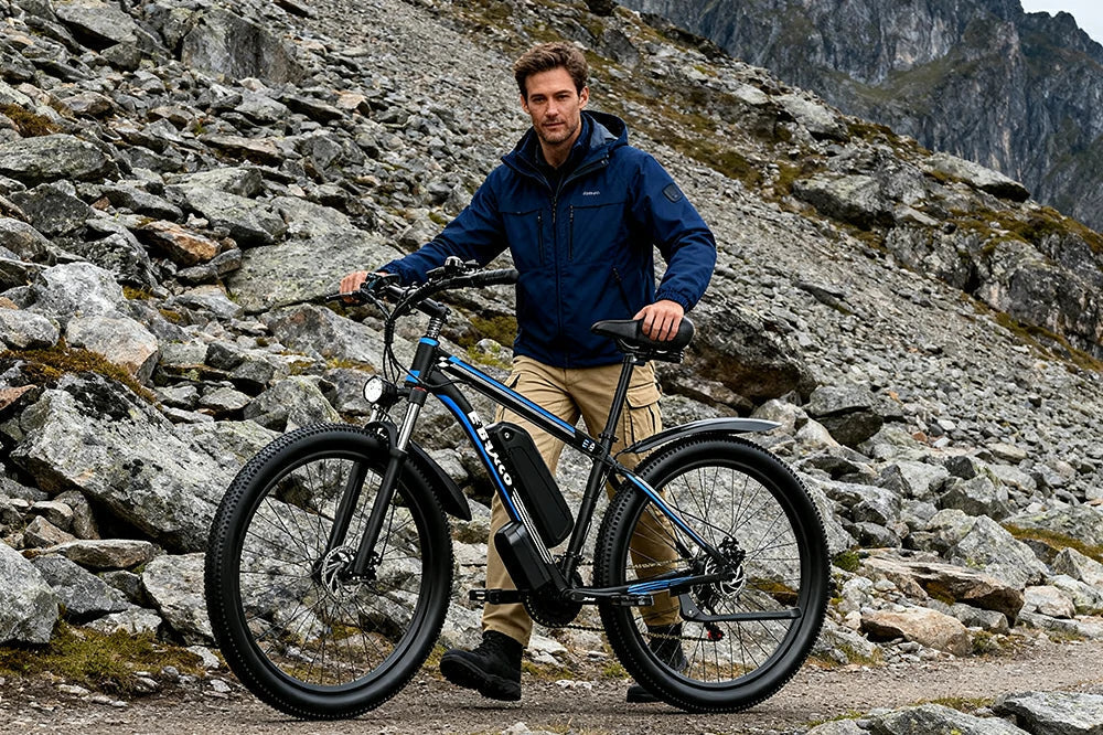 Alpine mountain lifestyle shot of a man standing beside black-blue E-BYCCO E8 electric mountain bike on rugged rocky mountain trail with jagged stone slopes in background.