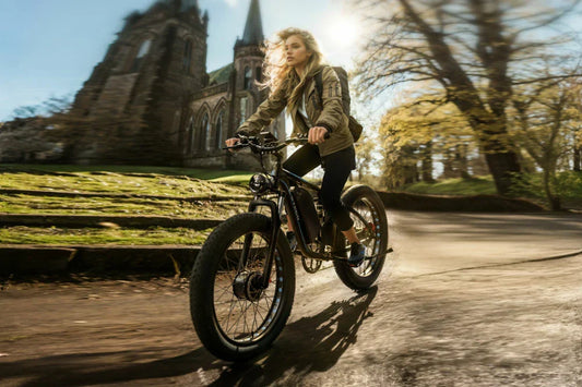 Matte black E-BYCCO EB7 Pro full suspension all-terrain electric bicycle, backpack-clad lady speeding on rural asphalt beside ancient stone cathedral and sunlit autumn woodland