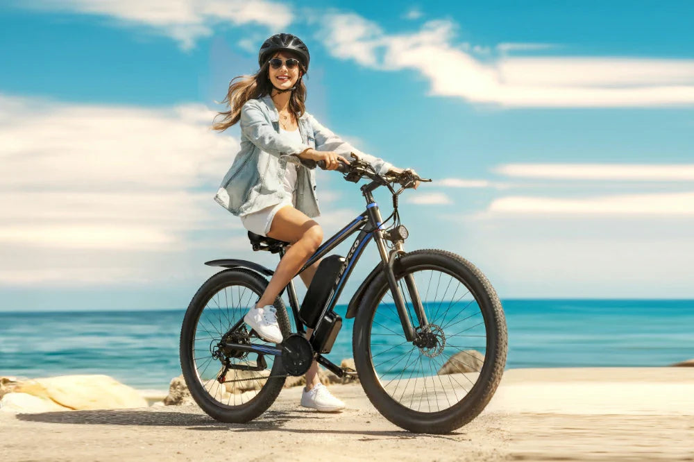 Bright coastal lifestyle shot of a woman riding black-blue E-BYCCO E8 electric mountain bike along sunny beach shore with turquoise ocean backdrop.